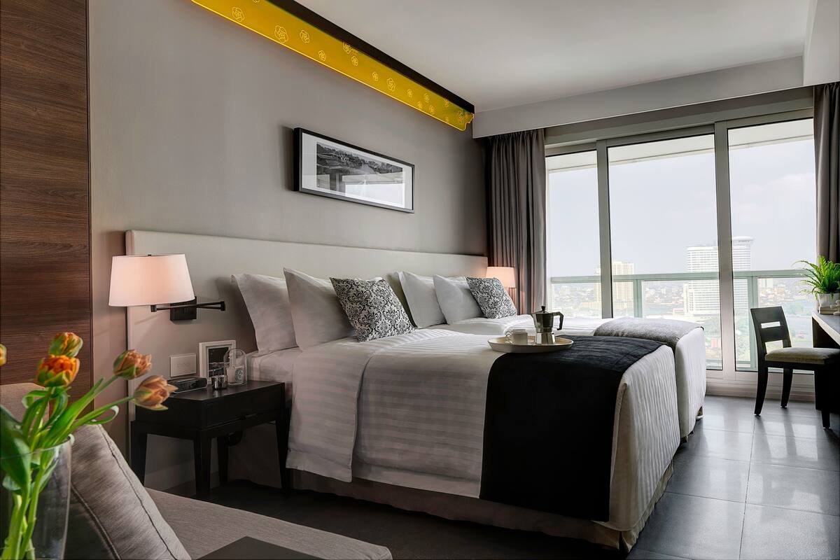 Une chambre d’hôtel moderne avec un grand lit, des lampes de chevet, une table basse, un fauteuil et une vue sur la ville grâce à de grandes fenêtres.