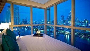 A hotel room with a large bed, a nightstand with a lamp, and a view of a city skyline at dusk. - Klapsons The River Residences Bangkok (Bangkok)