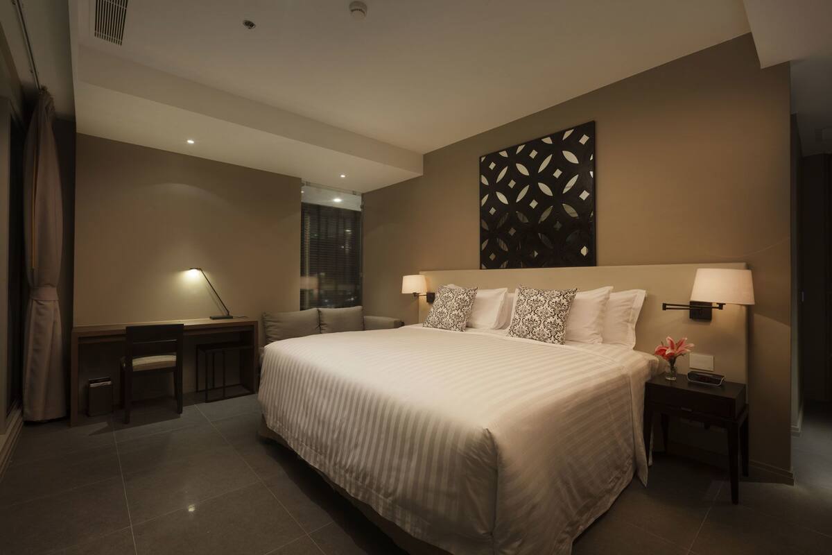 Une chambre d’hôtel moderne avec un grand lit, deux tables de chevet avec des lampes, un élément décoratif mural et une vue sur la ville par la fenêtre.