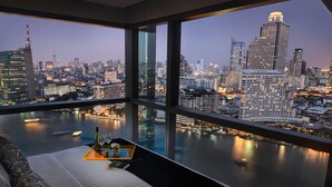 A high-rise hotel room with a panoramic city view, a bed, a tray with a bottle and glasses, and a table with a vase and flowers. - Klapsons The River Residences Bangkok (Bangkok)