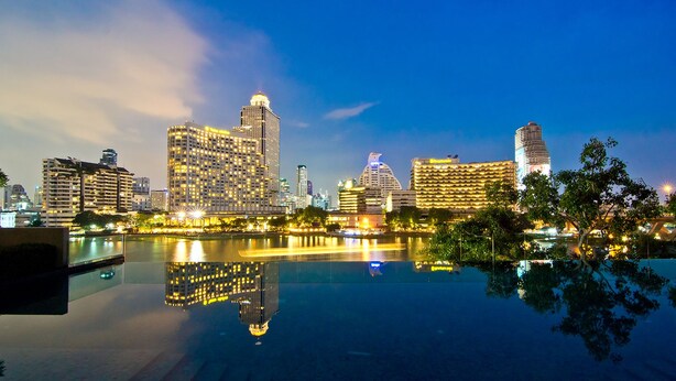 A city skyline at dusk with illuminated buildings reflecting on a calm body of water. - Klapsons The River Residences Bangkok (Bangkok)