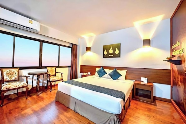 A hotel room with a large bed, a desk, two chairs, a nightstand, and a wall-mounted artwork. - The Ligor City Hotel (Nakhon Si Thammarat)