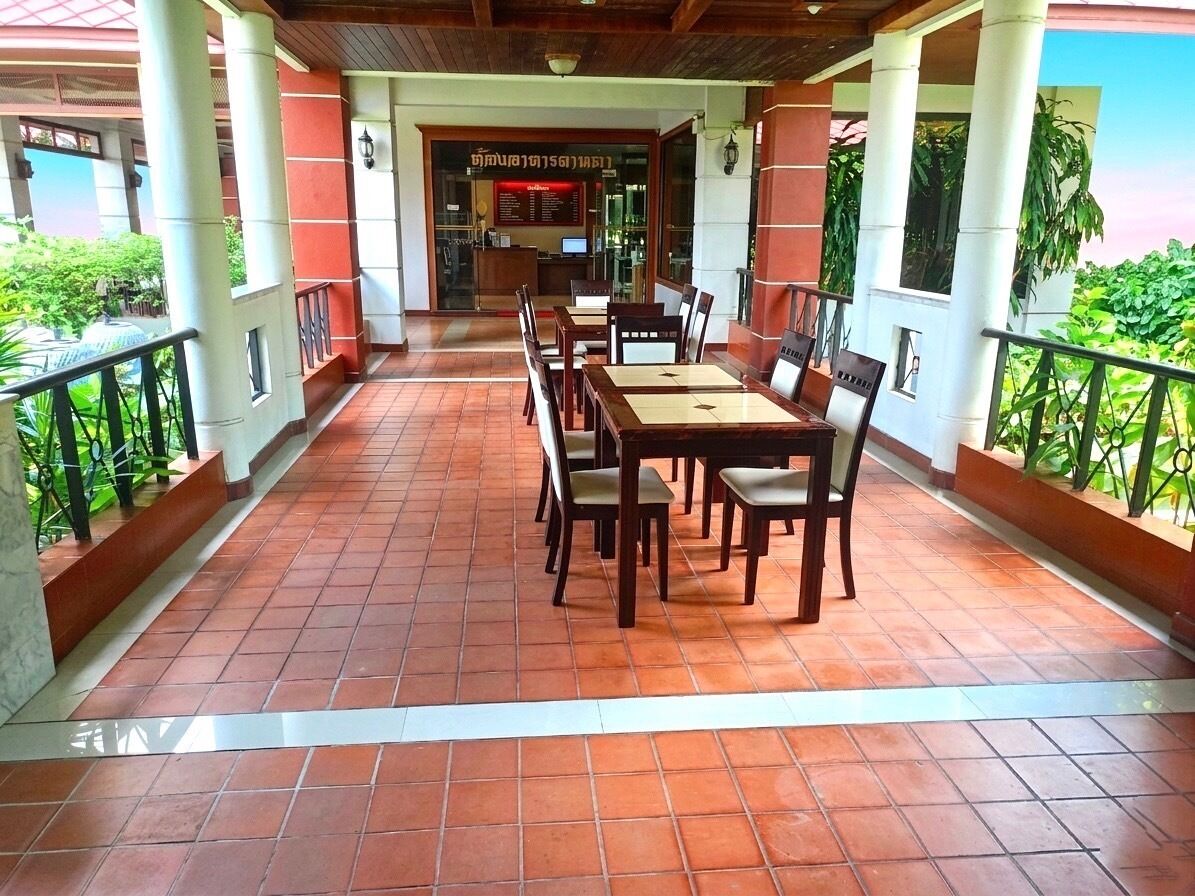 A covered outdoor dining area with a tiled floor, wooden tables and chairs, and a view of a restaurant in the background.