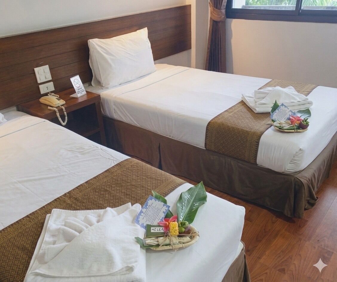 Two single beds with white linens, a bedside telephone, a 'No Smoking' sign, and a small table with a towel and a fruit basket.