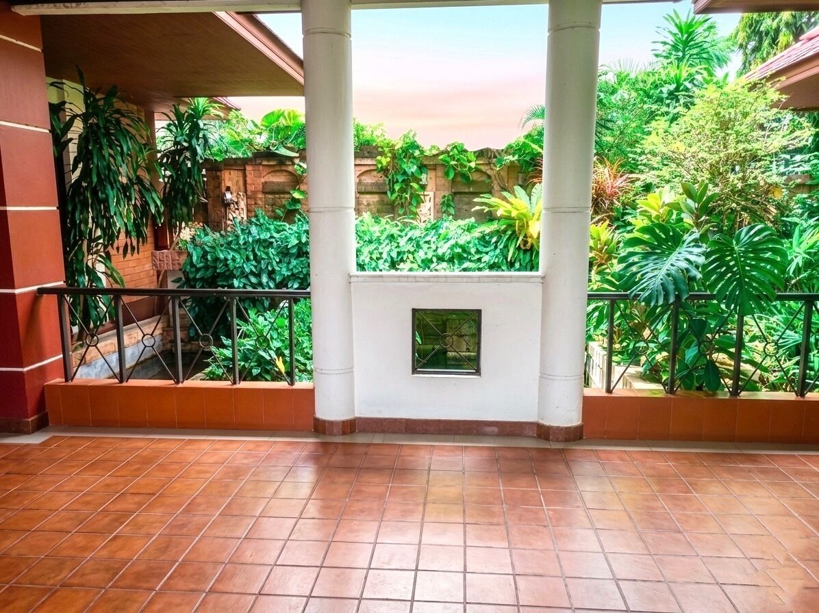 A covered balcony with tiled flooring, white pillars, and a view of lush greenery.