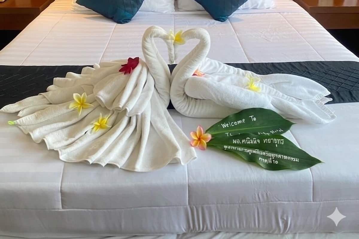 A hotel room with a neatly made bed, two pillows, a wall-mounted artwork, and a welcome sign with a flower.
