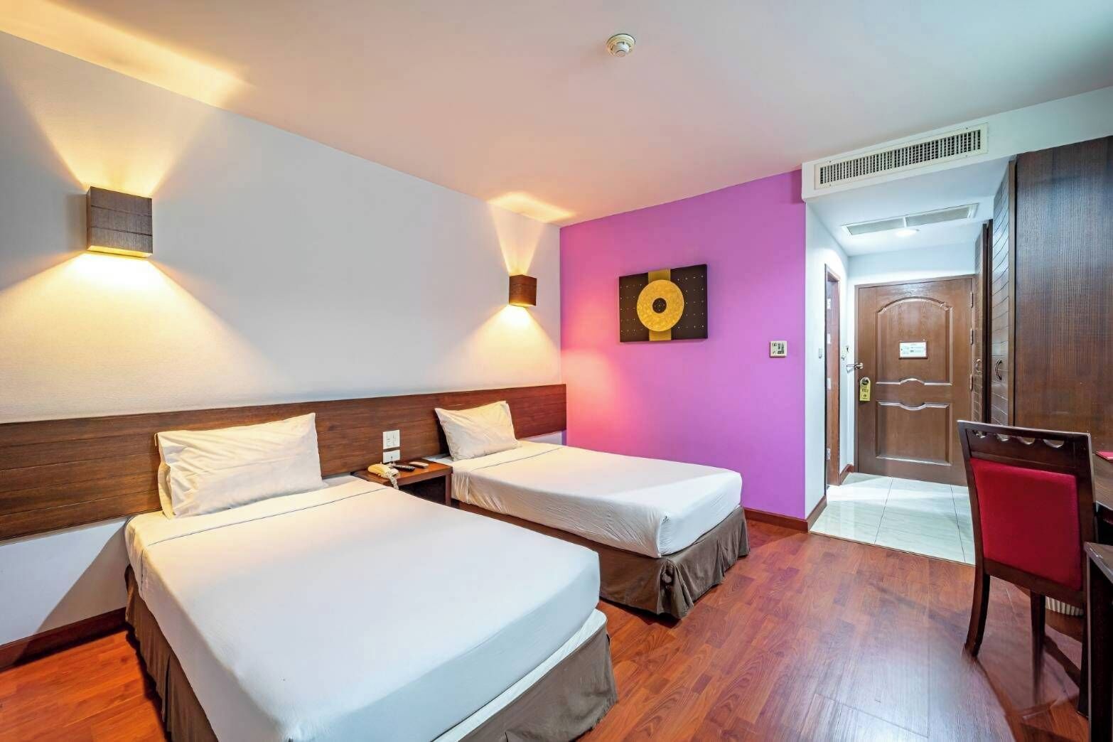 A hotel room with two beds, a wooden headboard, a wall-mounted light, a door, and a red chair.