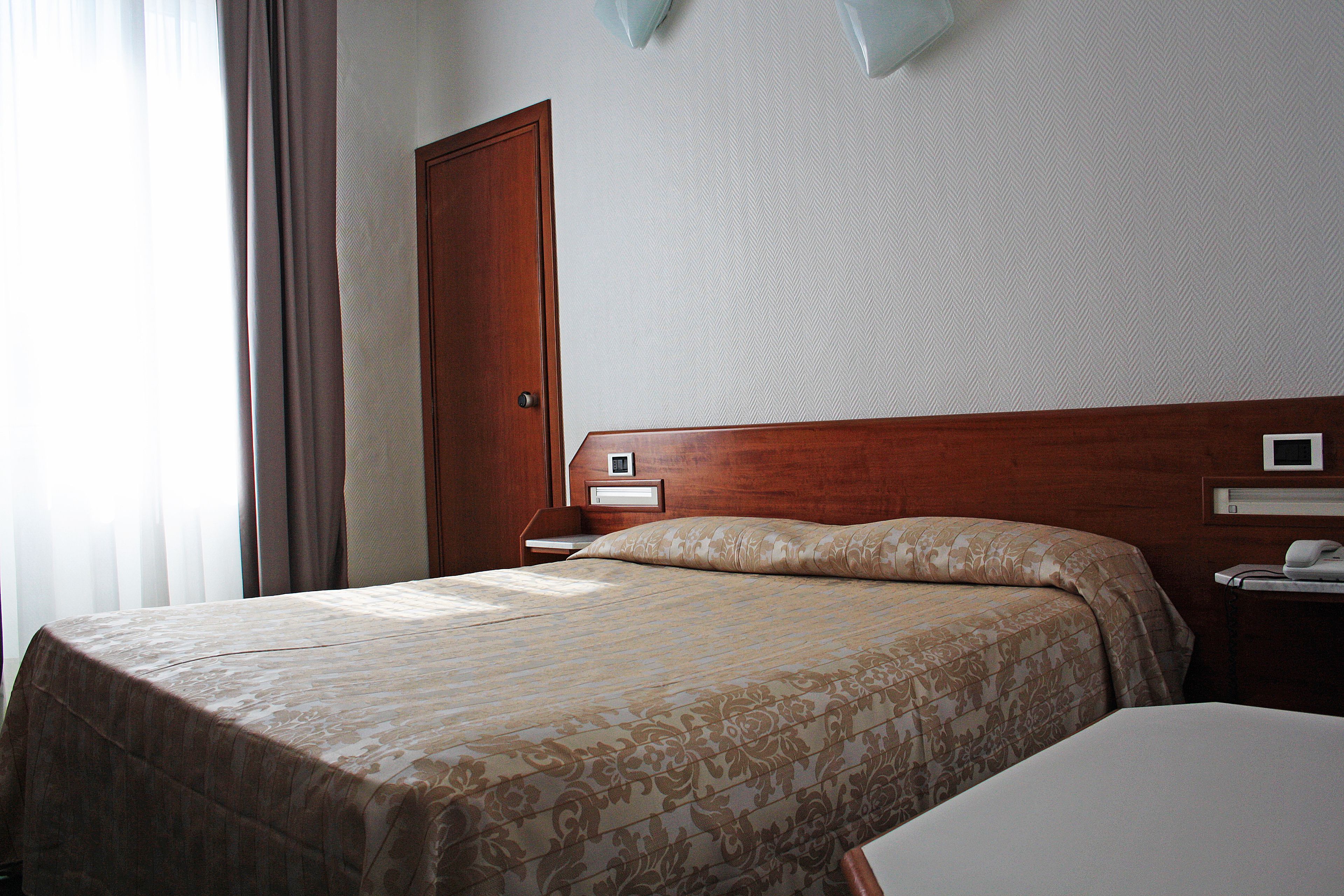 A hotel room with a wooden headboard, a bed with a patterned bedspread, a bedside table with a lamp, and a window with curtains.