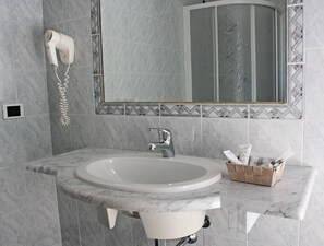 A bathroom with a marble sink, a hairdryer mounted on the wall, a mirror with a decorative frame, and a wicker basket on the countertop. - Hotel Centrale (Piombino)