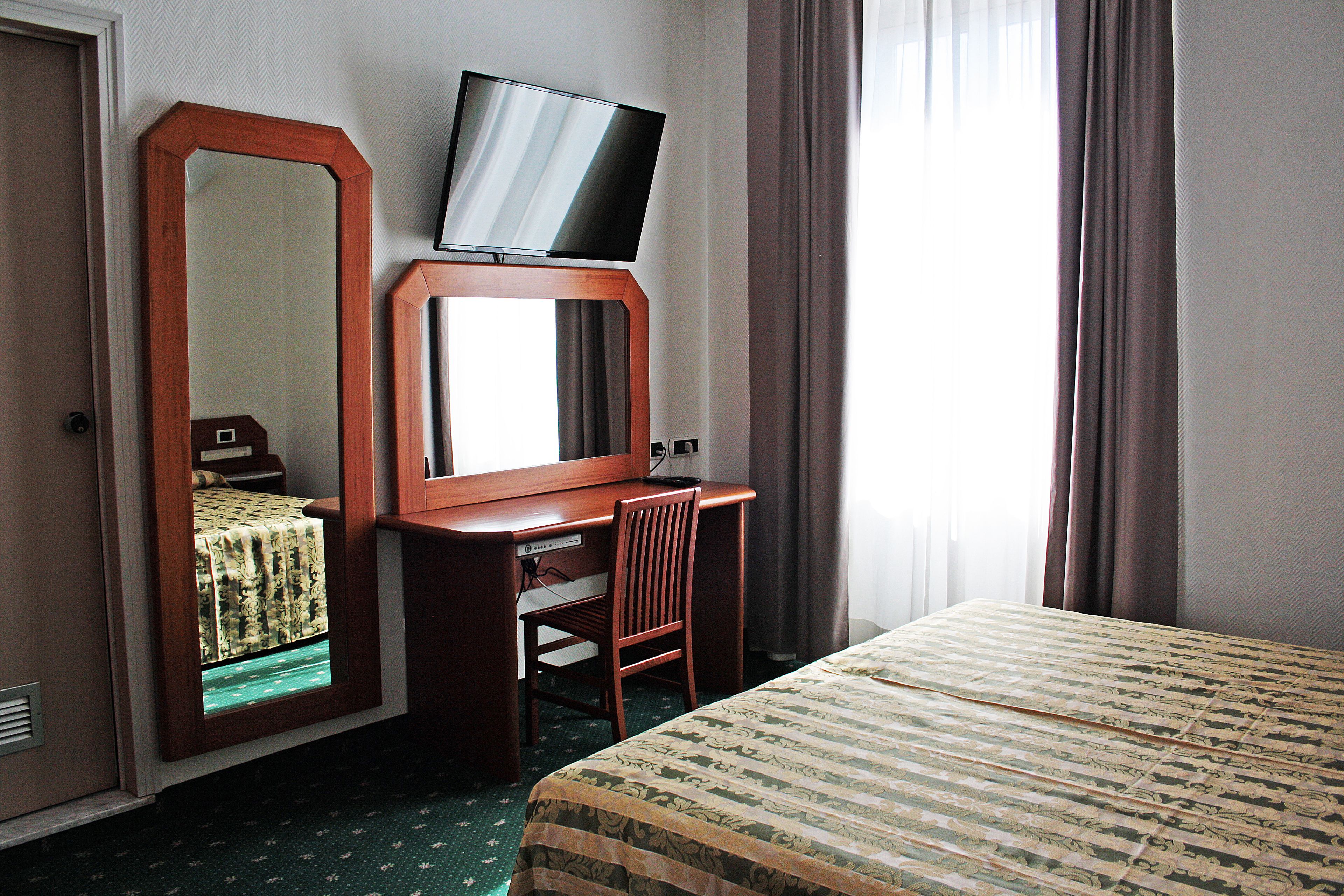 Ein Hotelzimmer mit Bett, Schreibtisch, Stuhl, Fernseher auf einem Standfuß und einem großen Spiegel.