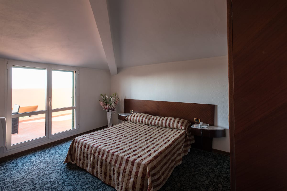 A hotel room with a large bed, a bedside table with a lamp, a window with a view, and a wooden headboard.