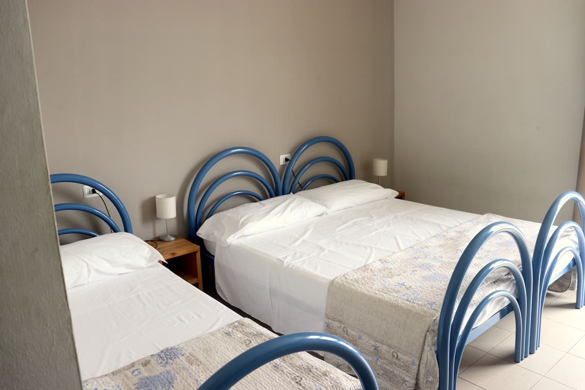 Ein Zimmer mit zwei Betten, jeweils mit blauem Metallgestell und weißer Bettwäsche. Dazwischen steht ein kleiner Nachttisch mit einer Lampe.