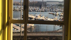 A view through a window of a marina with boats, a mountain in the background, and a building with a chimney. - Ostello del Porto (Lovere)