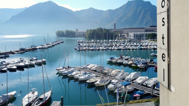 A marina with numerous boats docked, a mountain range in the background, and a building with the word "hostel" on it. - Ostello del Porto (Lovere)