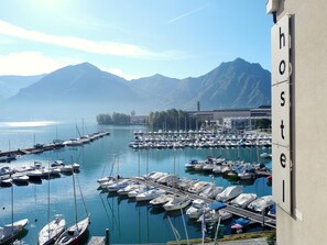 A marina with numerous boats docked, a mountain range in the background, and a building with the word "hostel" on it. - Ostello del Porto (Lovere)