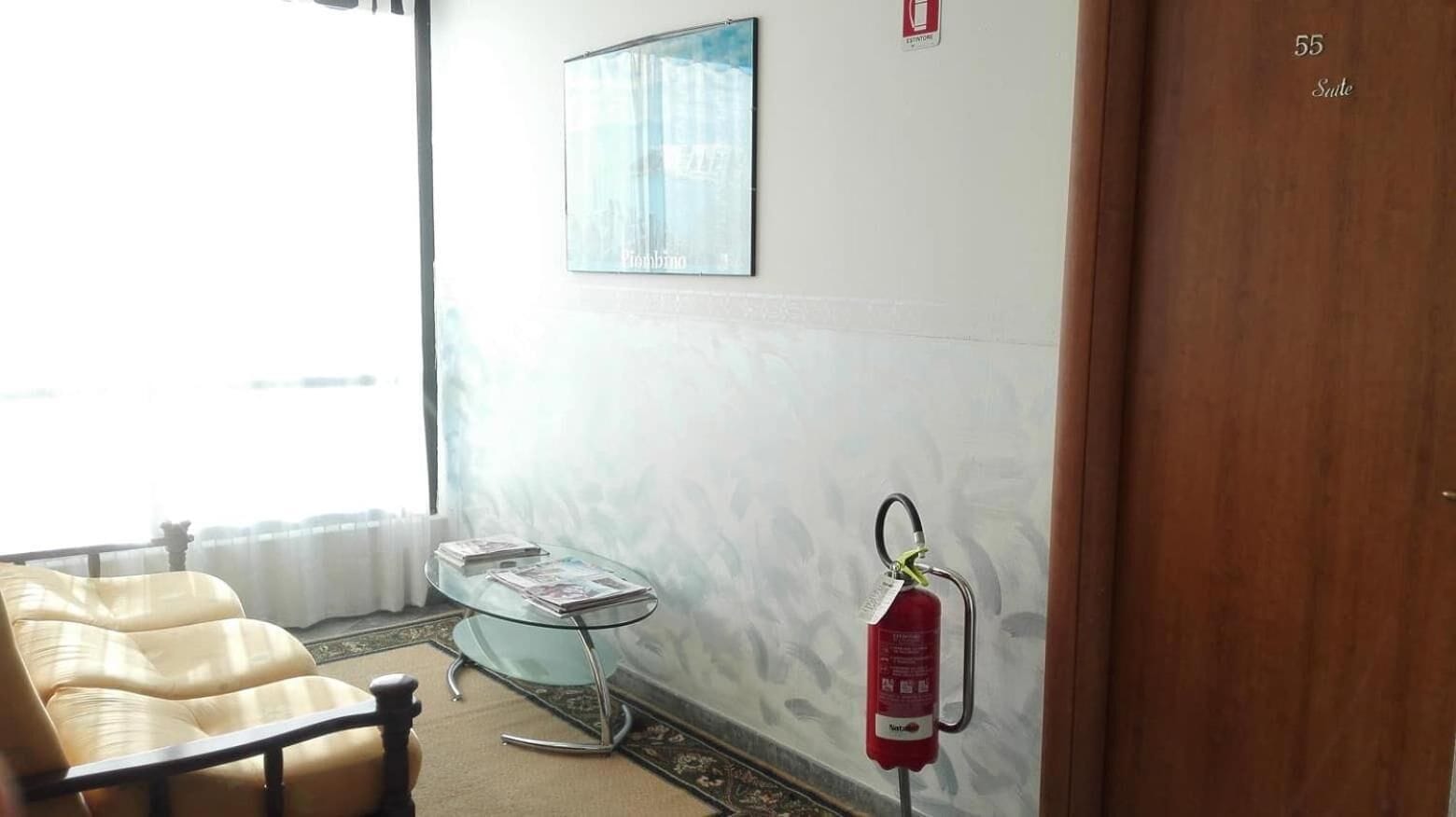 A hotel room with a sofa, a glass table with magazines, a fire extinguisher on the wall, and a door labeled "55 Suite".