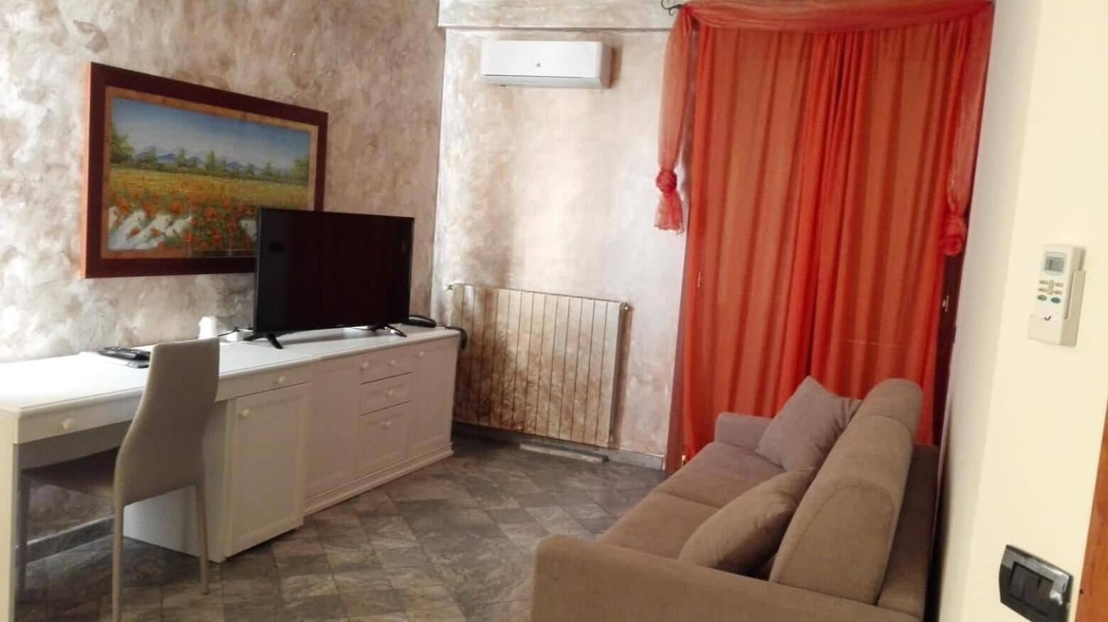 Ein Zimmer mit Schreibtisch, Fernseher, Sofa, Heizkörper und Vorhang.