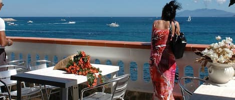 A terrace with tables and chairs, overlooking the sea with boats and a mountain in the distance.