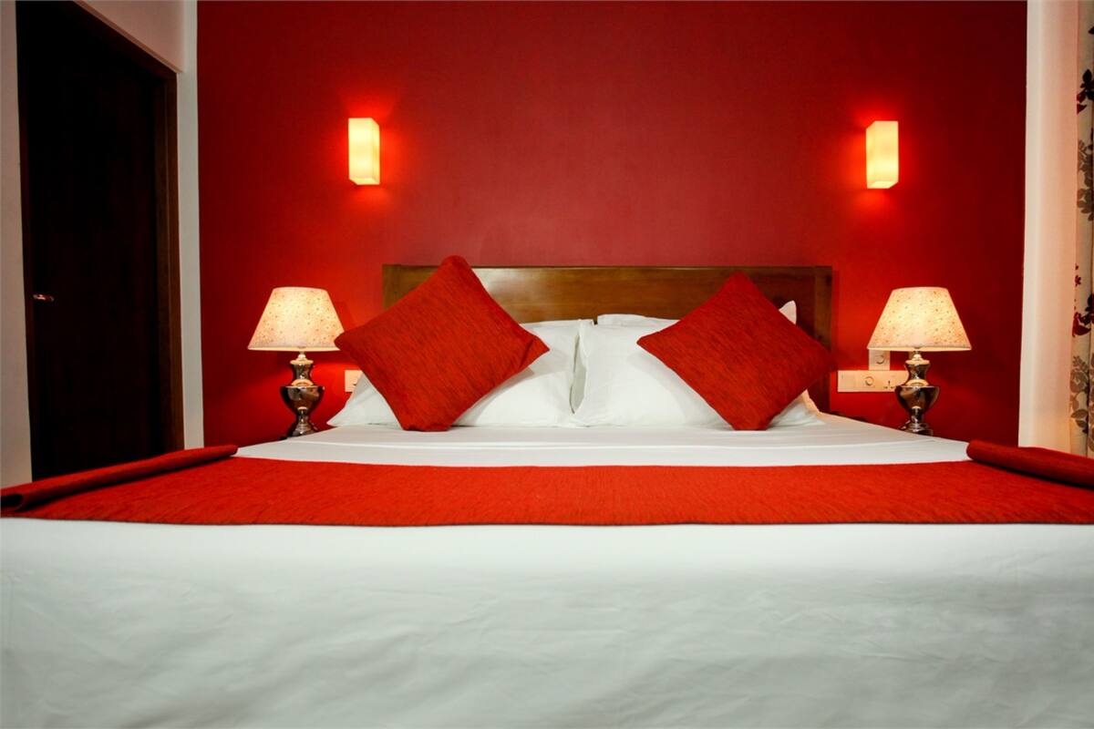 Habitación de hotel con alfombra roja, cabecera de madera, dos lámparas de noche y almohadas rojas.