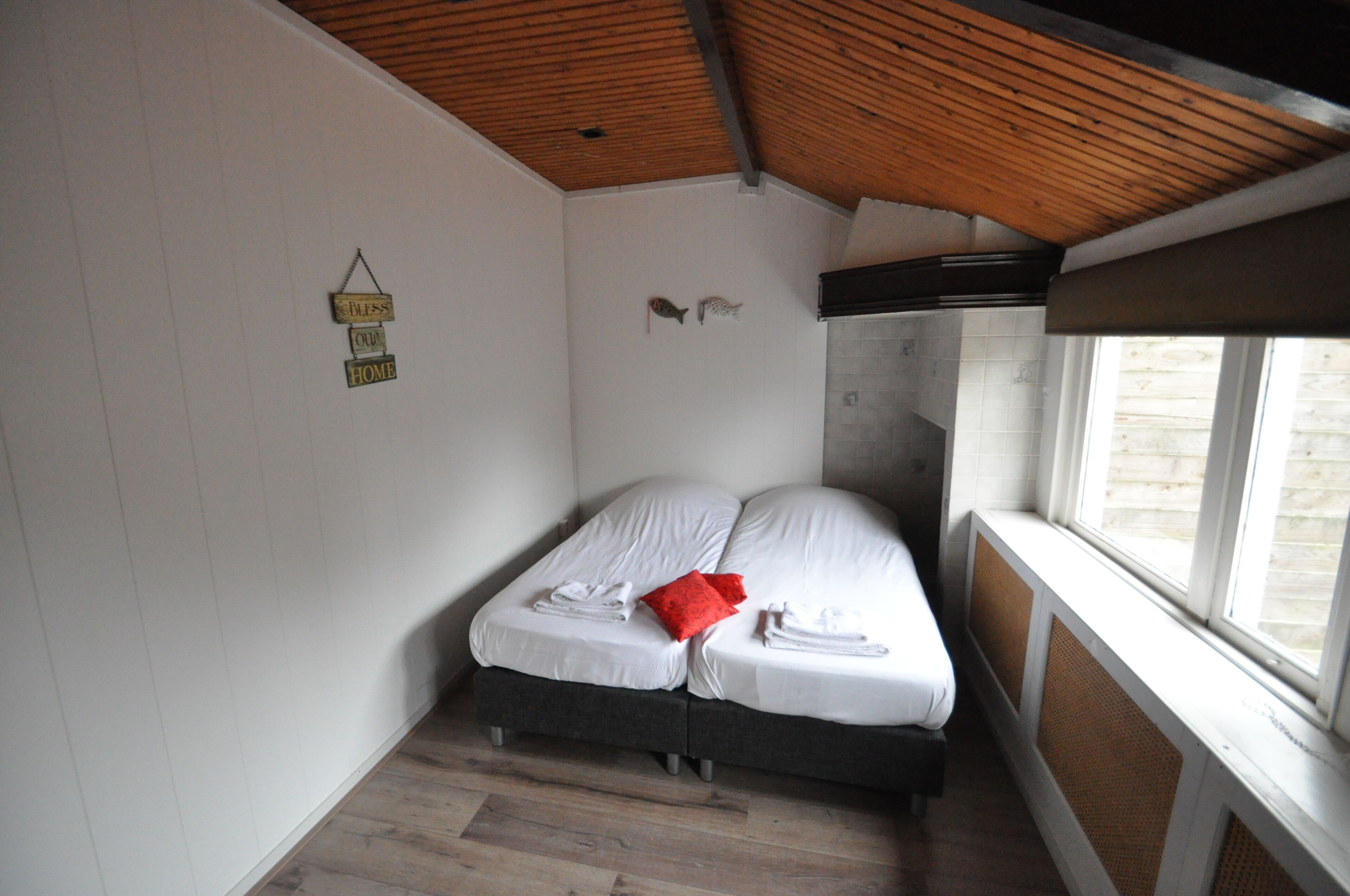 Ein Schlafzimmer mit Holzboden, zwei Betten mit weißen Bettwäsche, einem roten Kissen und einem kleinen Wandregal mit Dekoration.