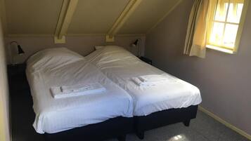 Un lit simple avec du linge de lit et des serviettes blanches, une petite table de chevet avec une lampe, et une fenêtre avec des rideaux.