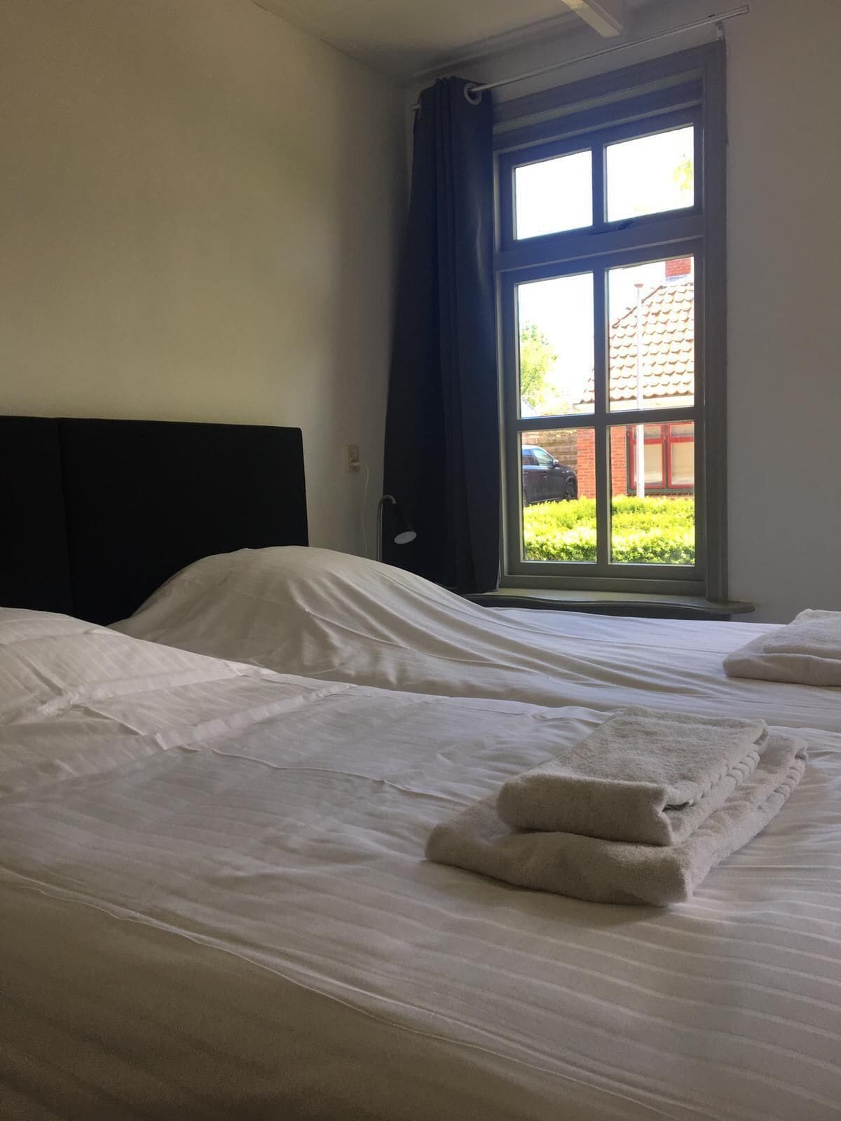 Une chambre d’hôtel avec deux lits, une grande fenêtre avec des rideaux et une vue sur un immeuble et de la verdure.
