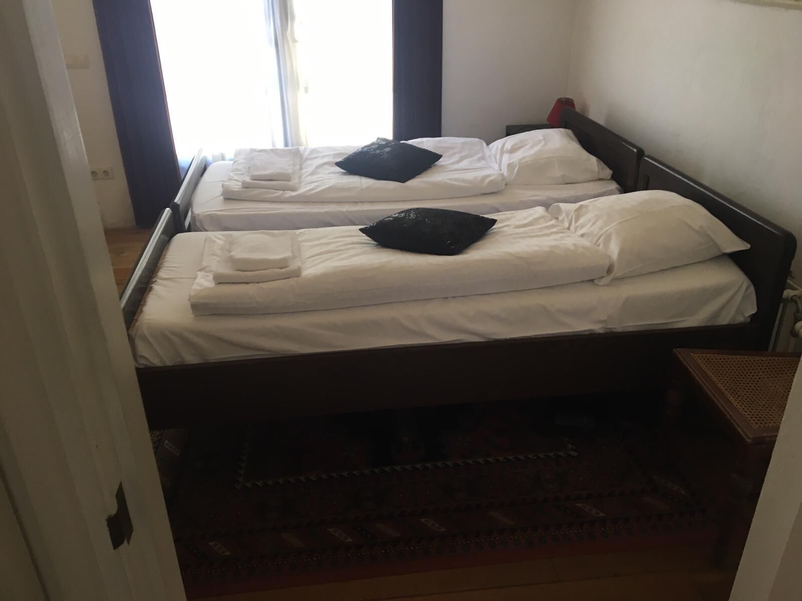 Ein Hotelzimmer mit zwei Einzelbetten, jeweils mit Kissen und gefaltetem Handtuch. Der Raum hat einen Holzboden und einen gemusterten Teppich.