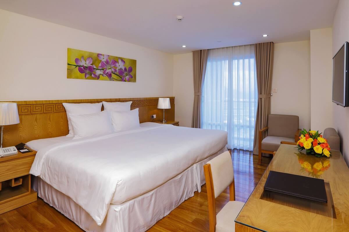 Een hotelkamer met een groot bed, een nachtkastje met een lamp, een bureau met een laptop, een stoel, een televisie en een schilderij aan de muur.