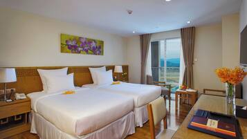 A hotel room with two beds, a small table with a vase of flowers, a desk with a telephone and a remote control, and a painting on the wall.
