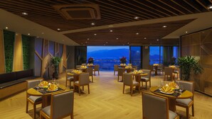 A modern dining area with wooden floors, round tables, and chairs. There are potted plants and a view of a cityscape through large windows. - Rosaka Nha Trang Hotel (Nha Trang)