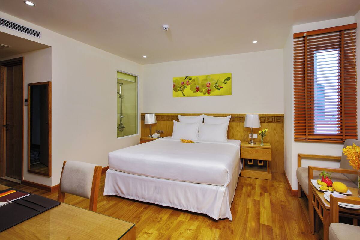 Een hotelkamer met een groot bed, een houten hoofdeinde, nachtkastjes met lampen, een kleintje tafeltje met een bloemenvaas, en een houten vloer.