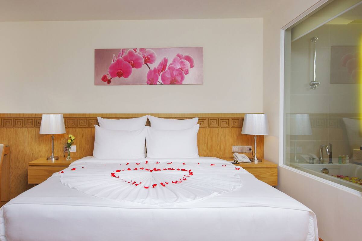 Een hotelkamer met een bed, een bureau, een stoel, een nachtkastje, een lamp, een vaas met bloemen en een schilderij aan de muur.