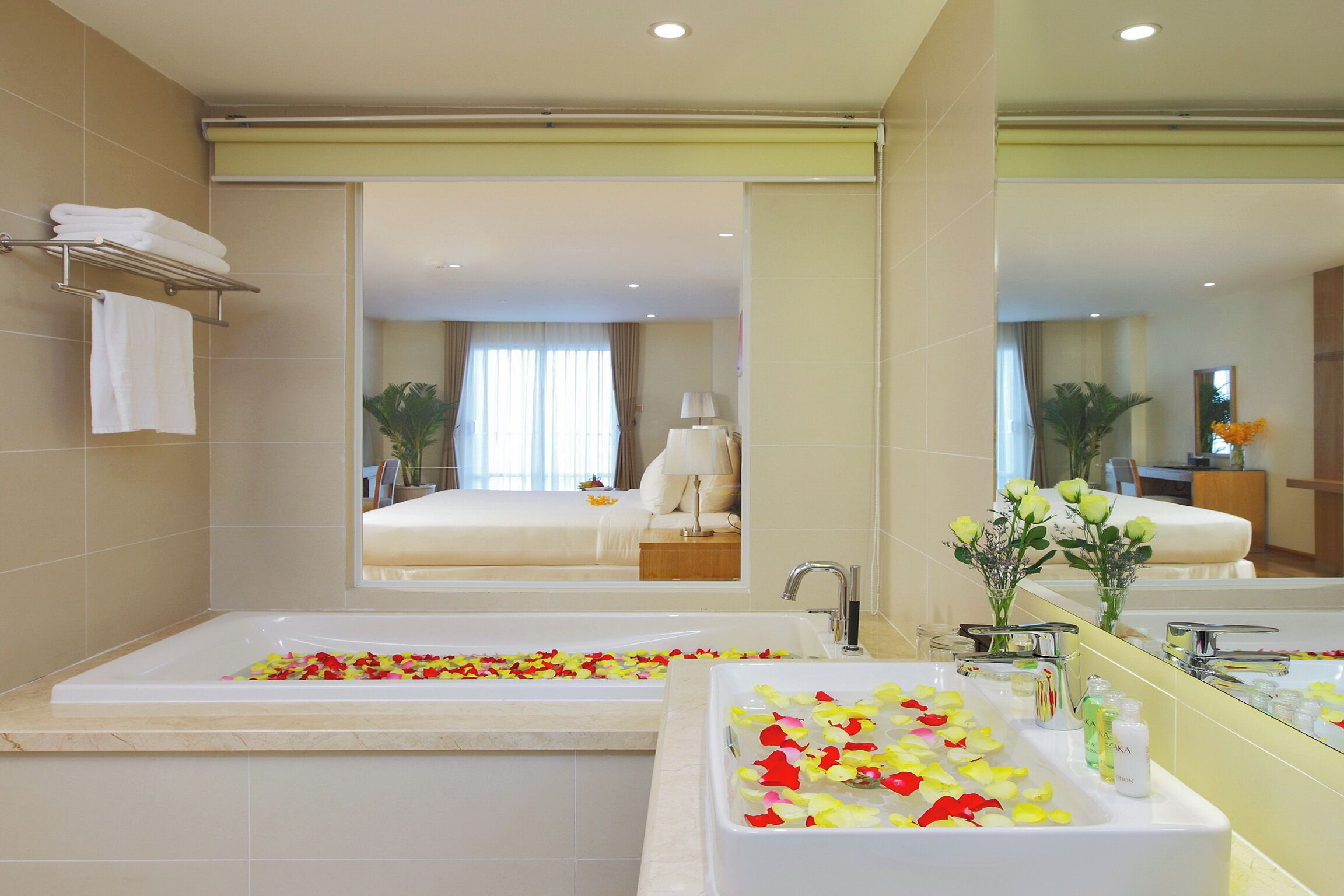 Une salle de bain moderne avec une baignoire et un lavabo, tous deux décorés de pétales de rose. Un grand miroir reflète la pièce, dans laquelle on aperçoit un lit et une table de chevet avec une lampe.