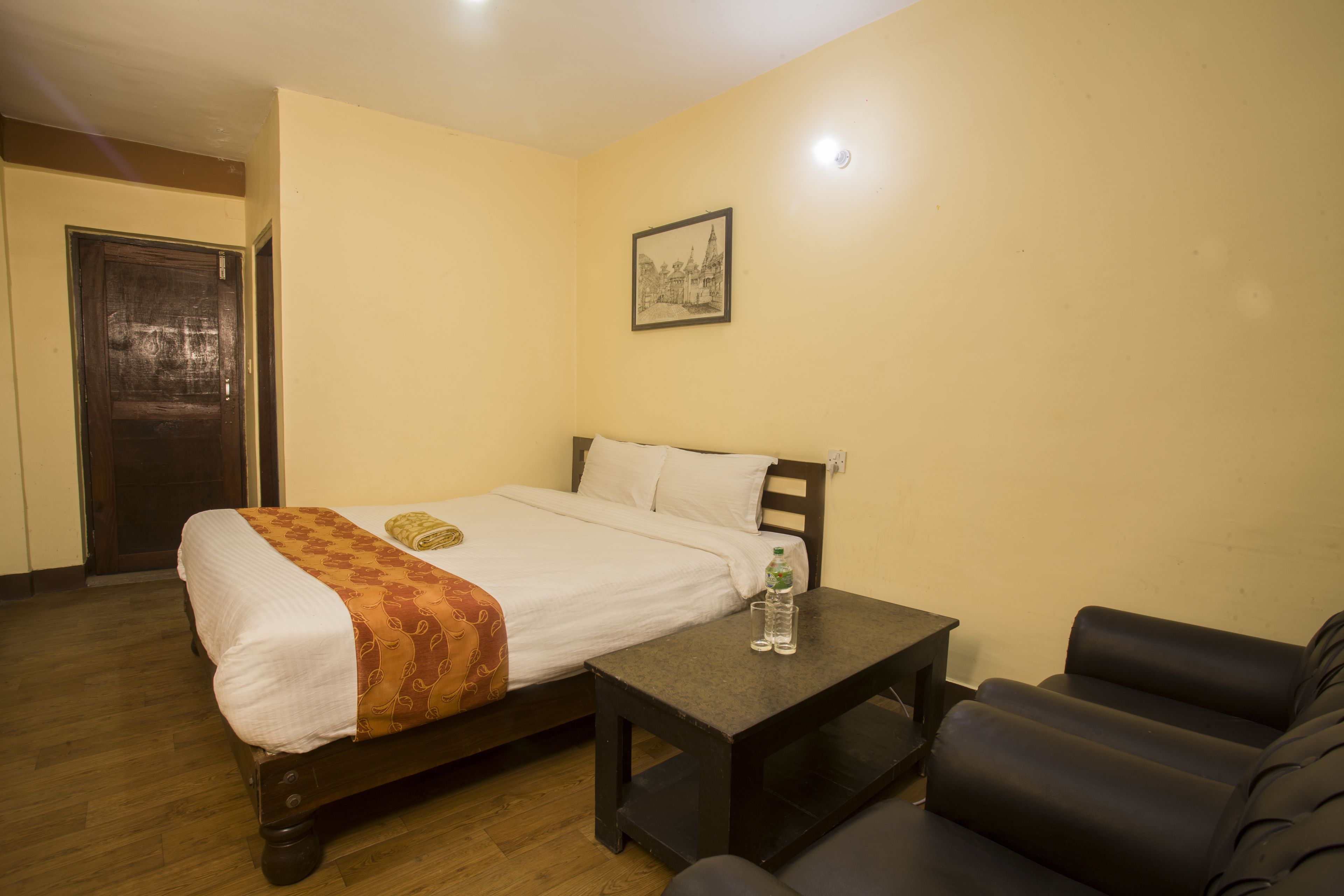 A single bed with a white and orange bedspread, a small table with a bottle, and a black leather couch in a room with wooden flooring and a single wall-mounted picture.