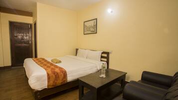 A single bed with a white and orange bedspread, a small table with a bottle, and a black leather couch in a room with wooden flooring and a single wall-mounted picture.