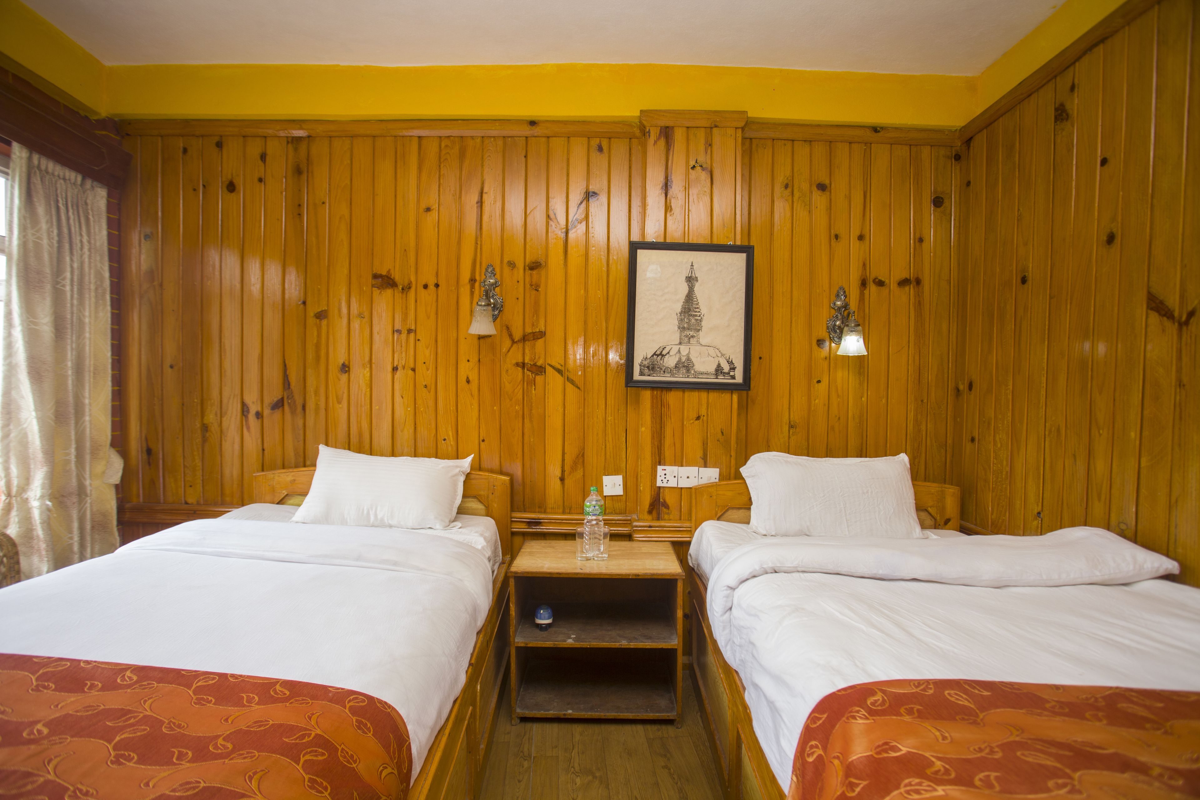 A hotel room with wooden paneled walls, two beds with white linens, a small wooden nightstand with a water bottle, and a framed picture on the wall.