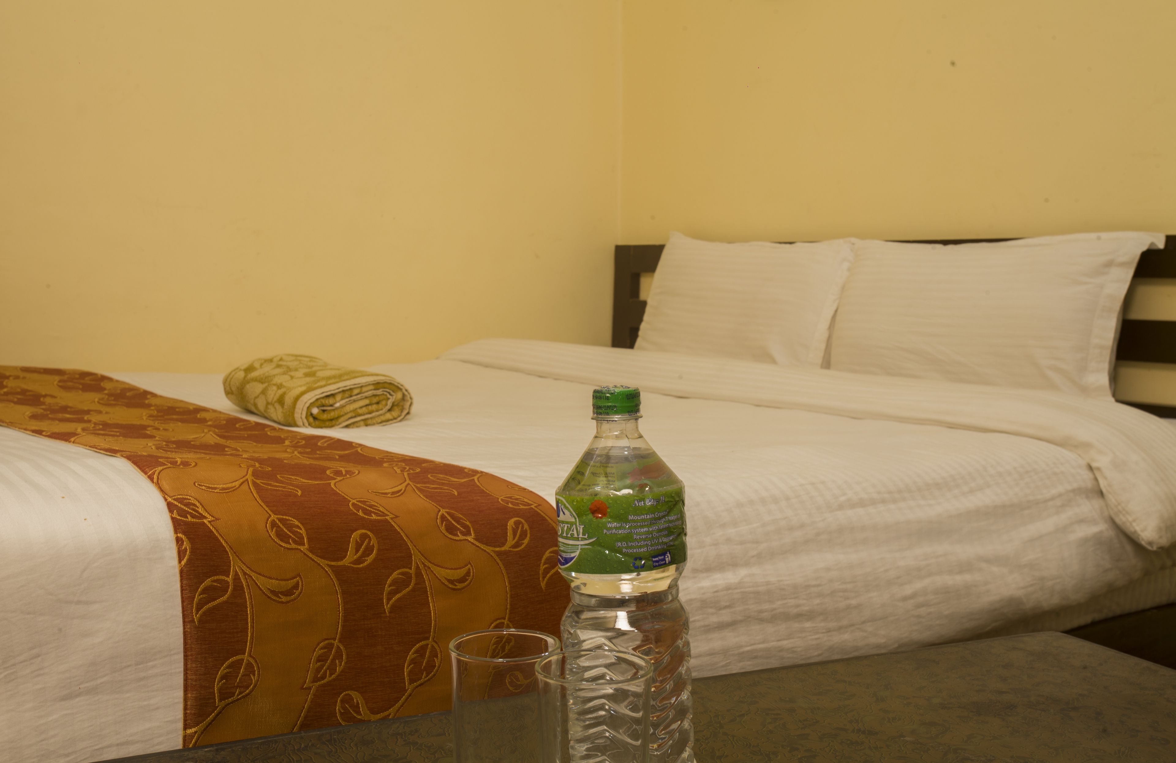 A hotel room with a bed, a rolled-up towel, a bottle of water, and a glass on a table.