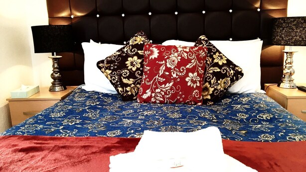 A bed with a blue patterned bedspread, a red blanket, and decorative pillows. There are two bedside tables with lamps and a tissue box on one. - Metro London City Airport (London)