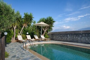 A pool area with lounge chairs and an umbrella, surrounded by trees and a stone wall. - Villa Athina (Chania)