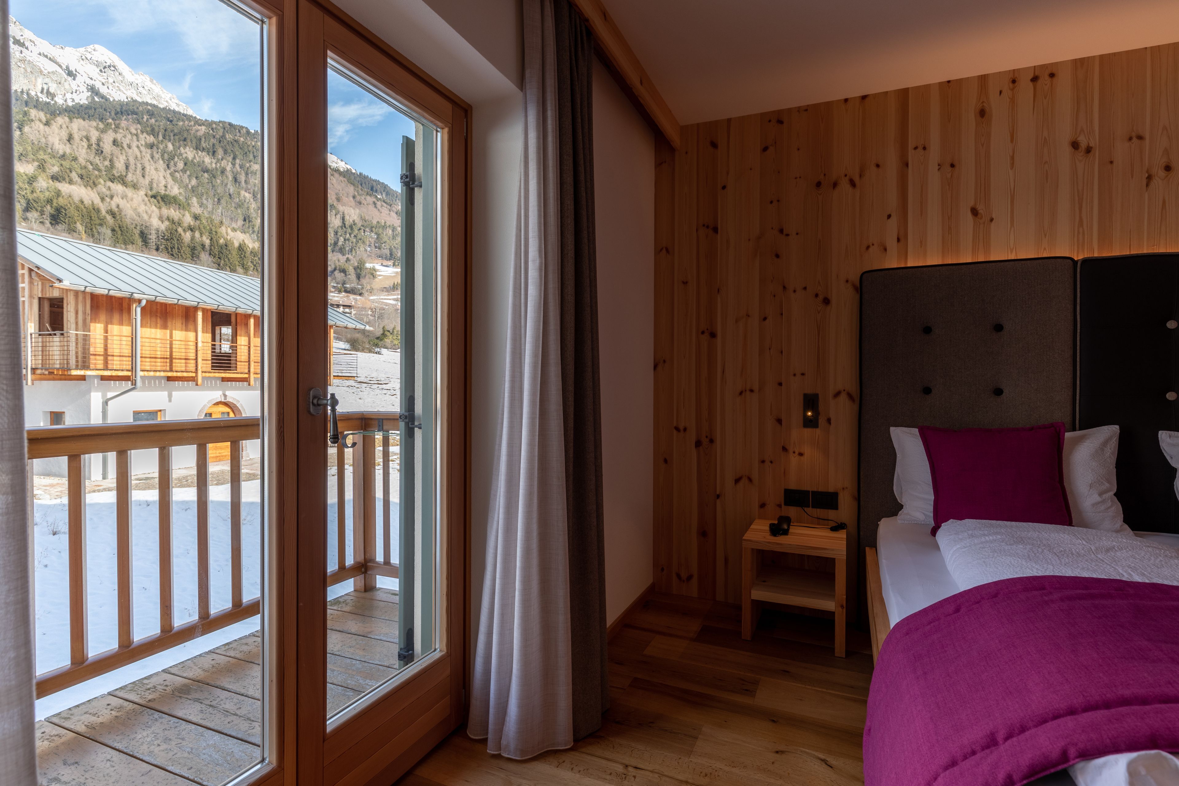 A bedroom with a large window, wooden walls, a bed with a headboard, and a small bedside table.