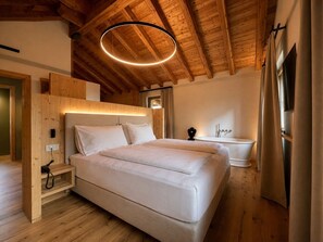 Quarto com teto de madeira, cama, mesa de cabeceira, luminária na parede e banheiro com banheira e cortinas.