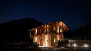 A two-story wooden house with a balcony, a stone wall, and a mountain in the background.