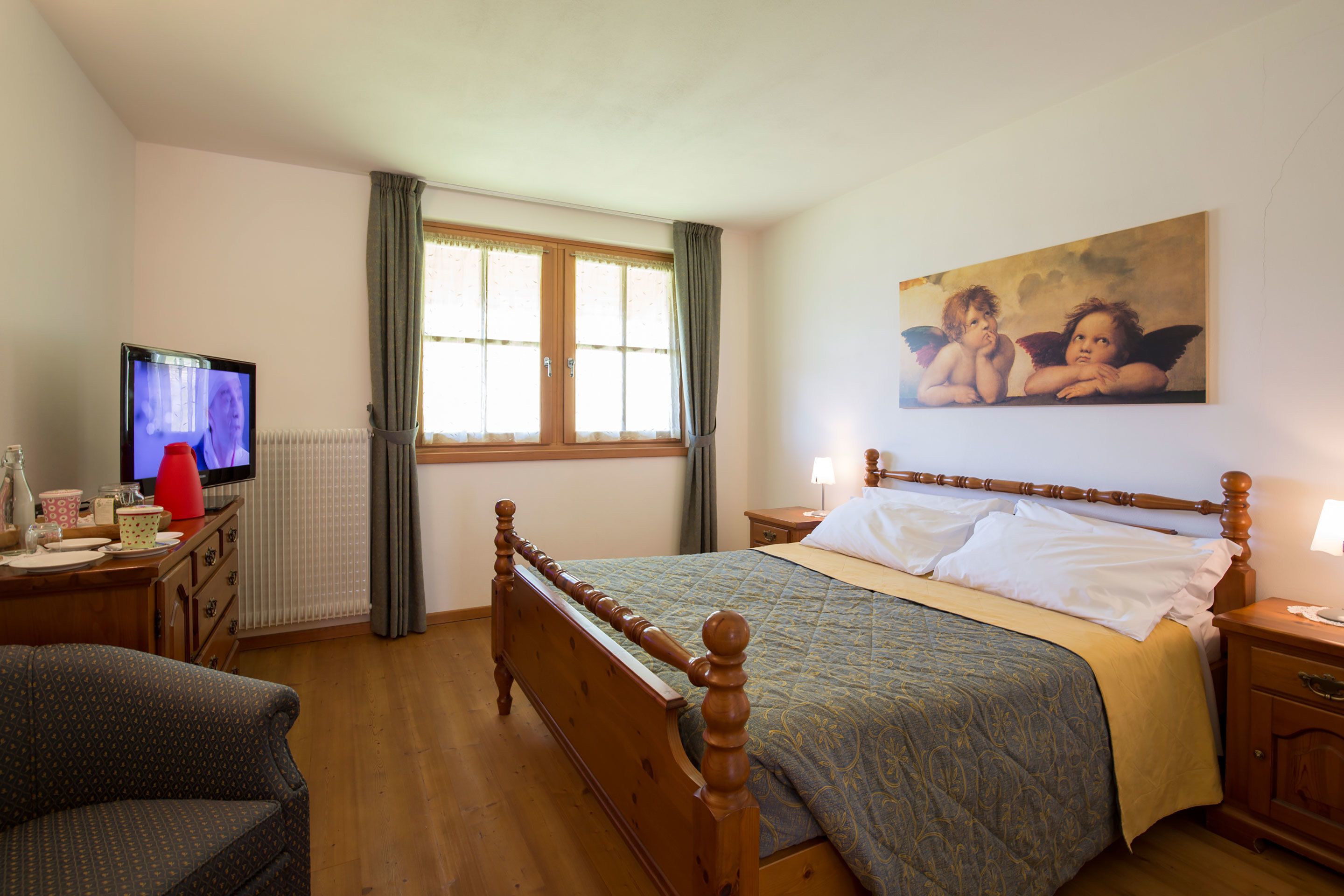 Une chambre à coucher avec un lit en bois, un téléviseur posé sur une commode, un tableau accroché au mur et une fenêtre avec des rideaux.