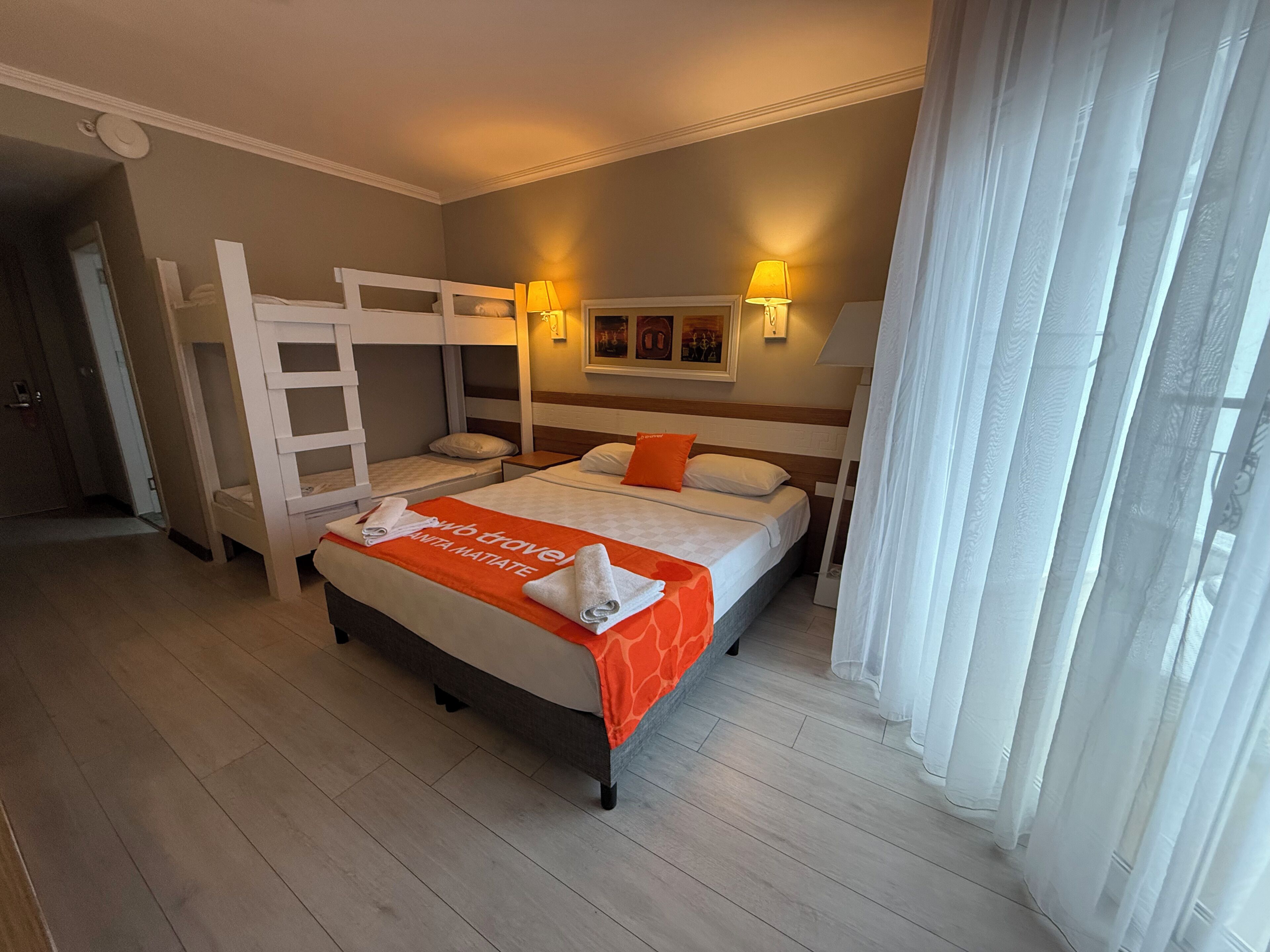 Ein Hotelzimmer mit einem Etagenbett, einem Einzelbett, einer Decke mit dem 'web travel'-Branding, zwei Nachttischlampen, einem Bild an der Wand und einem Fenster mit durchsichtigen Vorhängen.