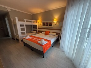 A hotel room with a bunk bed, a single bed, a 'web travel' branded blanket, two bedside lamps, a painting on the wall, and a window with sheer curtains. - Anita Matiate Hotel - All inclusive (Kemer)