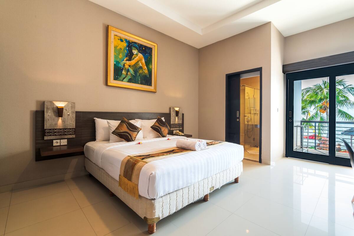 A spacious bedroom with a large bed, a nightstand, a painting, and a door leading to a balcony with a view of palm trees.