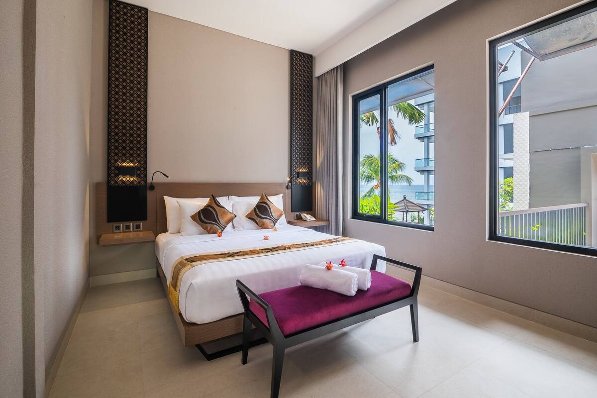 A hotel room with a bed, a bench, a TV, and a window with a view of palm trees and a building.