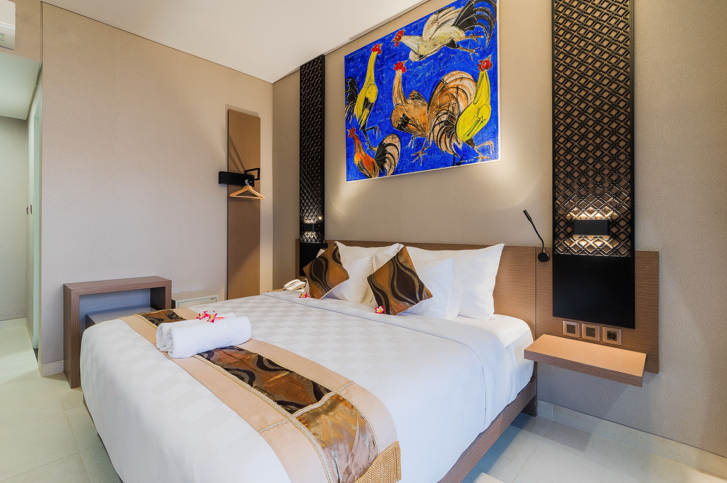 A modern hotel room with a large bed, a painting of roosters, a small desk, and a wall-mounted TV.
