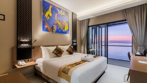 Ein Hotelzimmer mit einem großen Bett, einem Hahn-Gemälde, einem Balkon mit Sonnenuntergangsblick und einem Schreibtisch mit Stuhl.