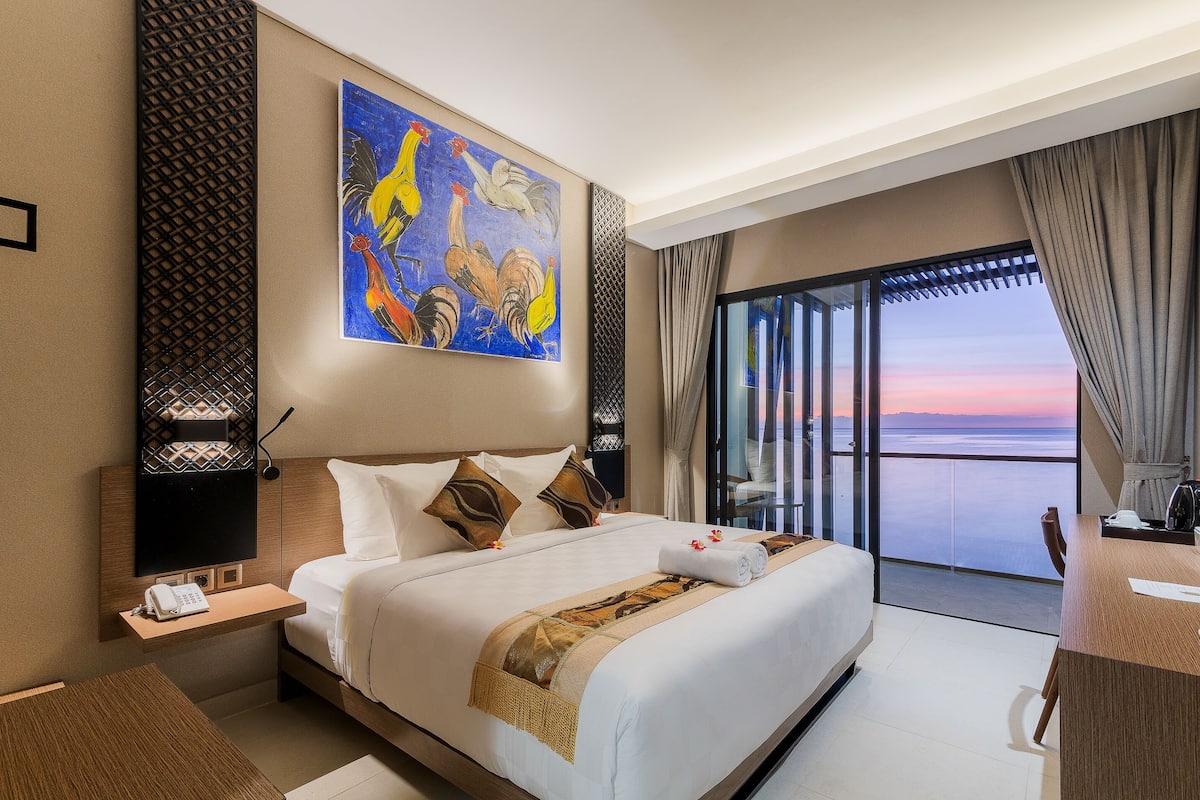 A hotel room with a large bed, a painting of roosters, a balcony with a view of the sunset, and a desk with a chair.